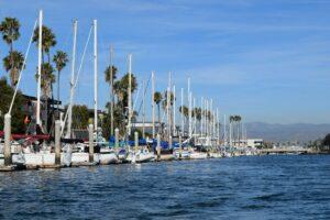 A serene view of the marina in Oxnard, California, showcasing a lineup of sailboats moored neatly under a clear blue sky, with palm trees and distant mountains completing the picturesque coastal scene.